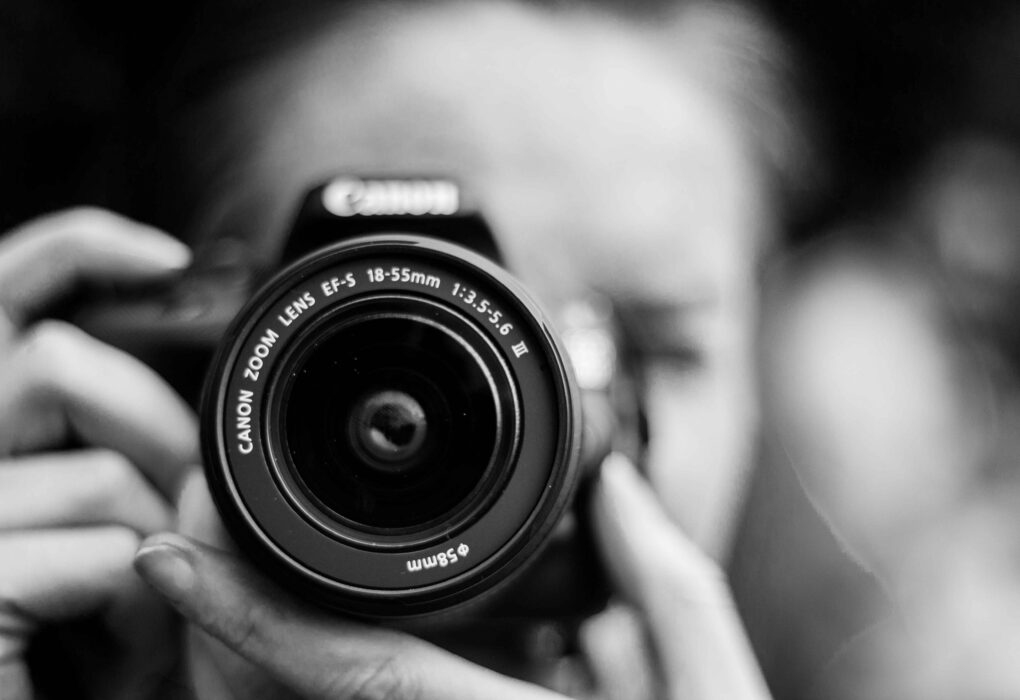 Close-up of a person holding a Canon DSLR camera with 18-55mm zoom lens, black and white photography