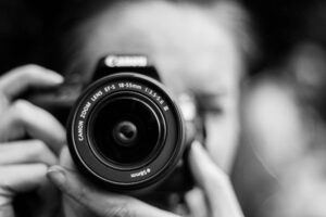 Close-up of a person holding a Canon DSLR camera with 18-55mm zoom lens, black and white photography